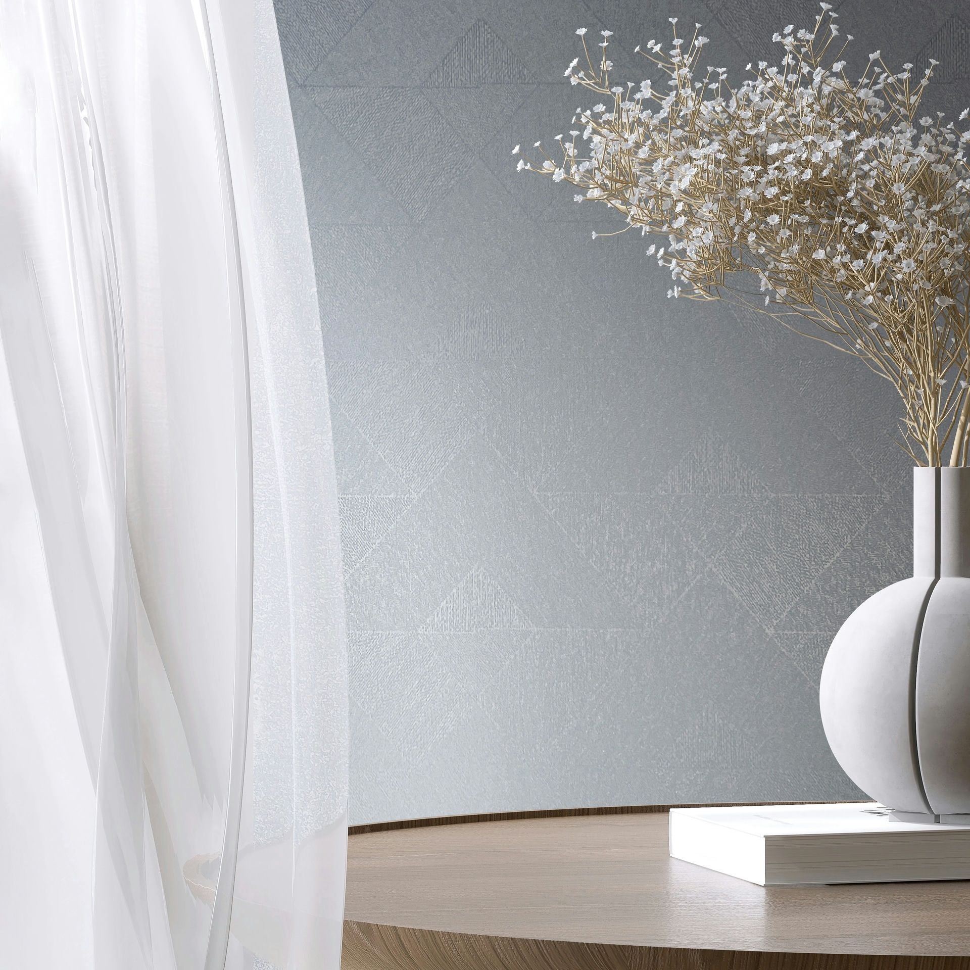 Marburg Naomi 82547 Styled room scene with blue‑gray wallpaper featuring a subtle geometric triangle pattern behind a sheer white curtain, a round wooden table, a sculptural ceramic vase holding delicate dried flowers, and a closed book.