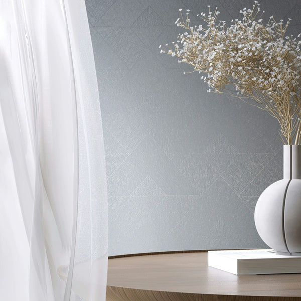 Marburg Naomi 82547 Styled room scene with blue‑gray wallpaper featuring a subtle geometric triangle pattern behind a sheer white curtain, a round wooden table, a sculptural ceramic vase holding delicate dried flowers, and a closed book.