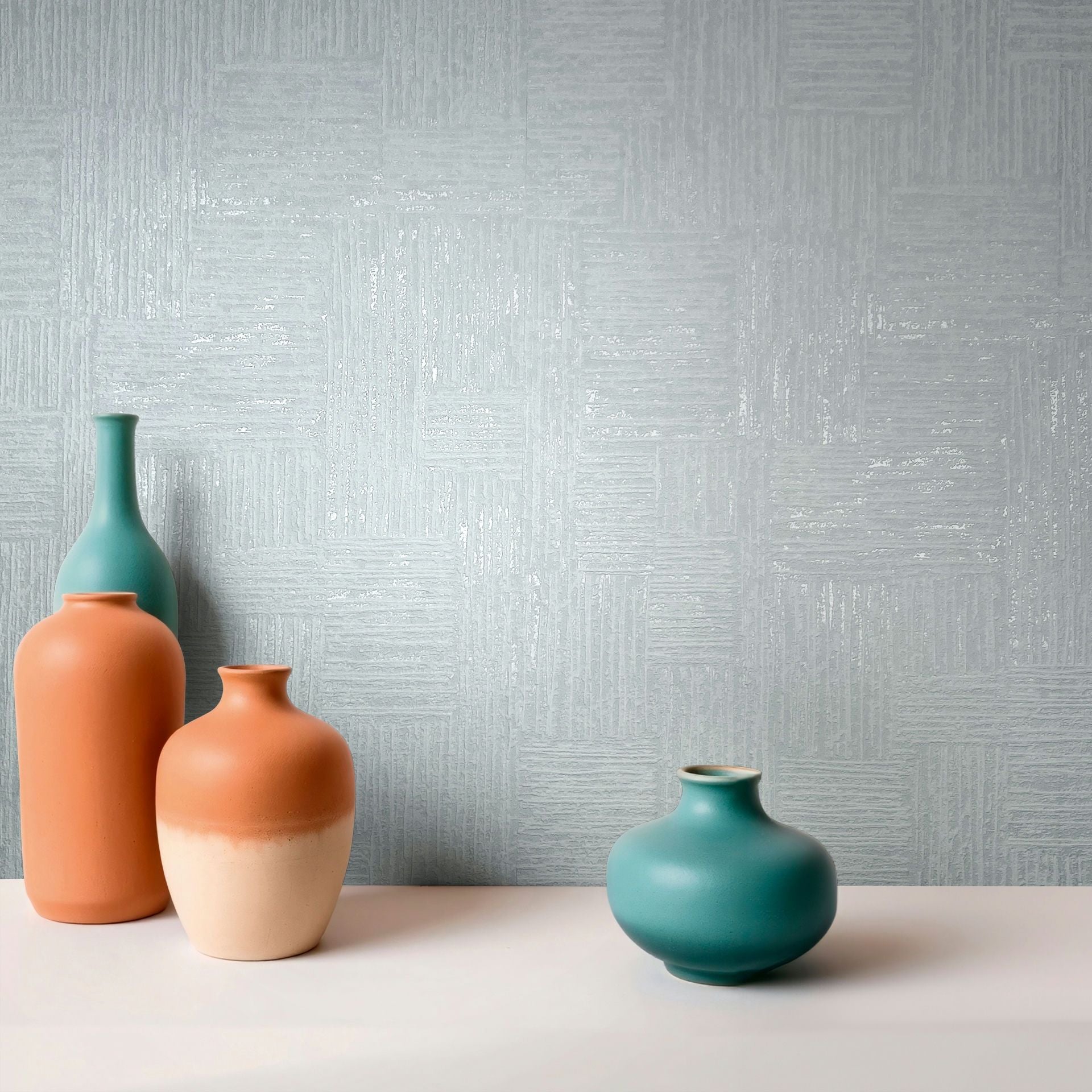 Marburg Naomi 82551 Light blue wallpaper with a subtle geometric block pattern and slight sheen, shown behind four teal and terracotta ceramic vases on a light surface.