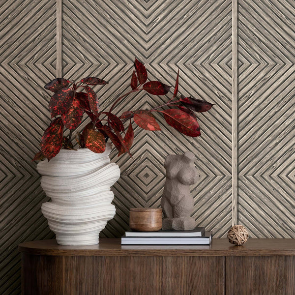Marburg Naomi 89018 Beige and gray geometric diamond wallpaper behind a wooden console styled with a white sculptural vase holding red foliage, a small torso sculpture, stacked books, a small wooden container, and a rattan ball.