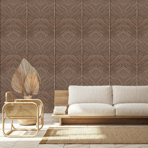 Marburg Naomi 89020 Brown geometric diamond-line wallpaper creates a feature wall behind a light upholstered sofa, a rattan chair, a small table holding dried palm leaves in a pot, a woven rug, and a stack of books.