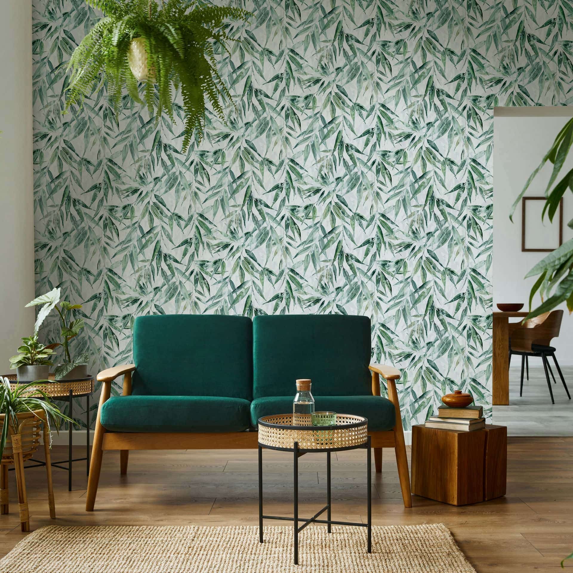 Marburg Naomi 89039 Living room with a dense green leafy wallpaper on a light background behind a green sofa, a round cane-topped side table, a wood block side table, potted plants, and a doorway to a dining area.
