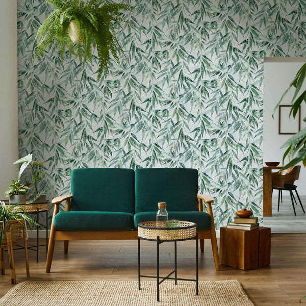 Marburg Naomi 89039 Living room with a dense green leafy wallpaper on a light background behind a green sofa, a round cane-topped side table, a wood block side table, potted plants, and a doorway to a dining area.