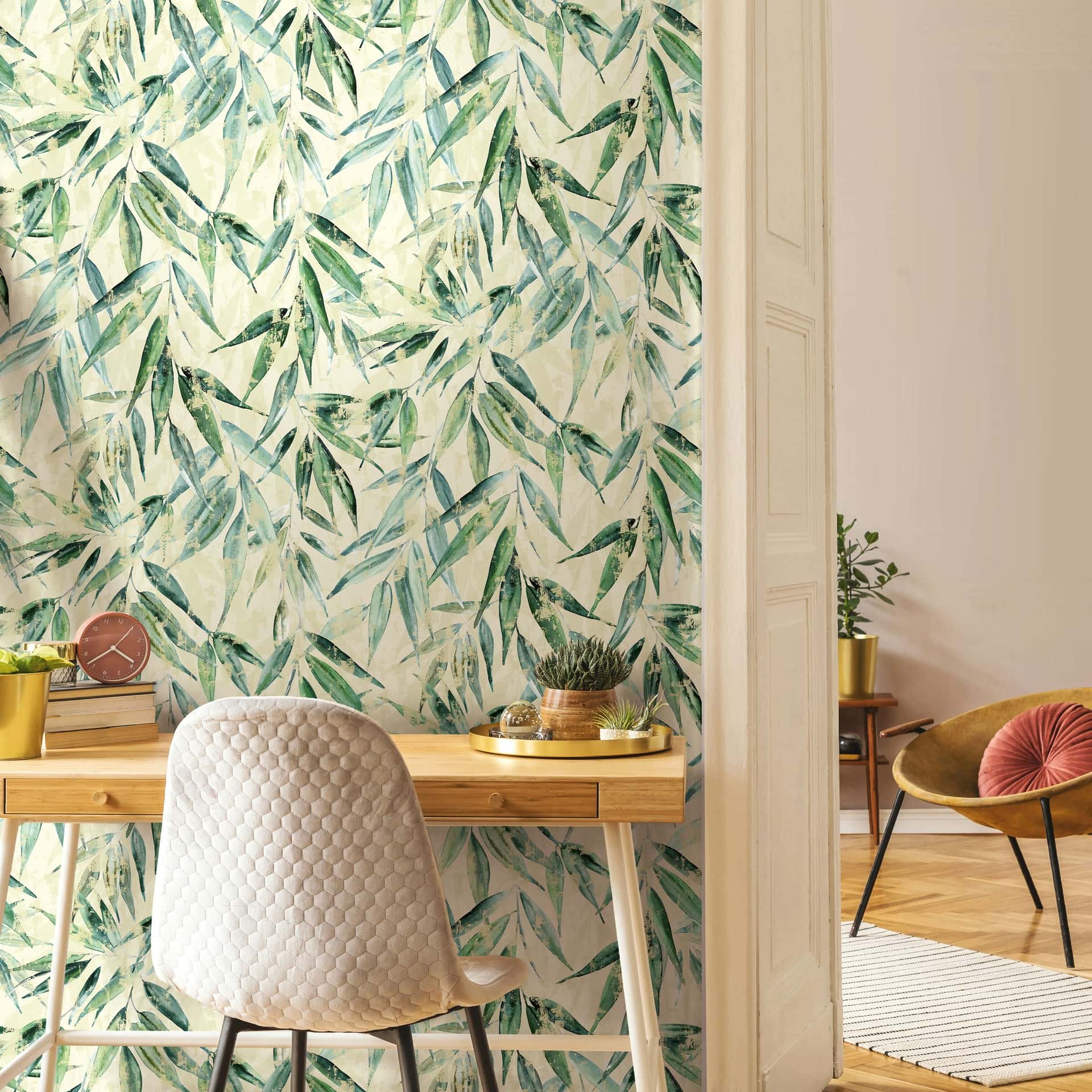 Marburg Naomi 89040 Leafy green botanical wallpaper on a light background behind a wooden desk with a quilted chair; small plants and books on the desk and a glimpse of a lounge chair and rug in the adjacent area.