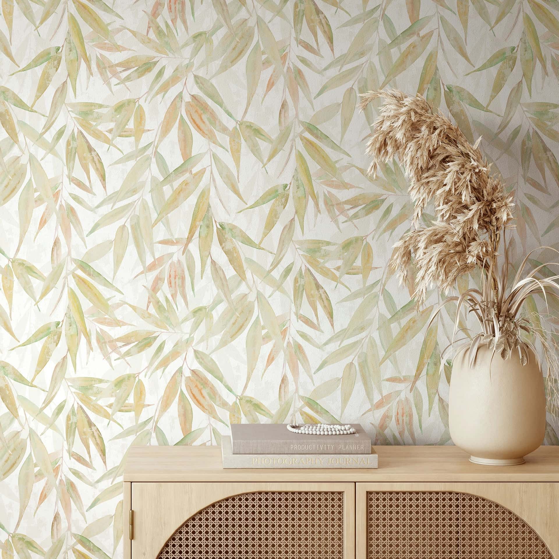 Marburg Naomi 89042 Leaf-pattern wallpaper with watercolor-style slender leaves in green and warm tones on a light background, shown behind a light wood console with a ceramic vase of dried grasses and stacked books.