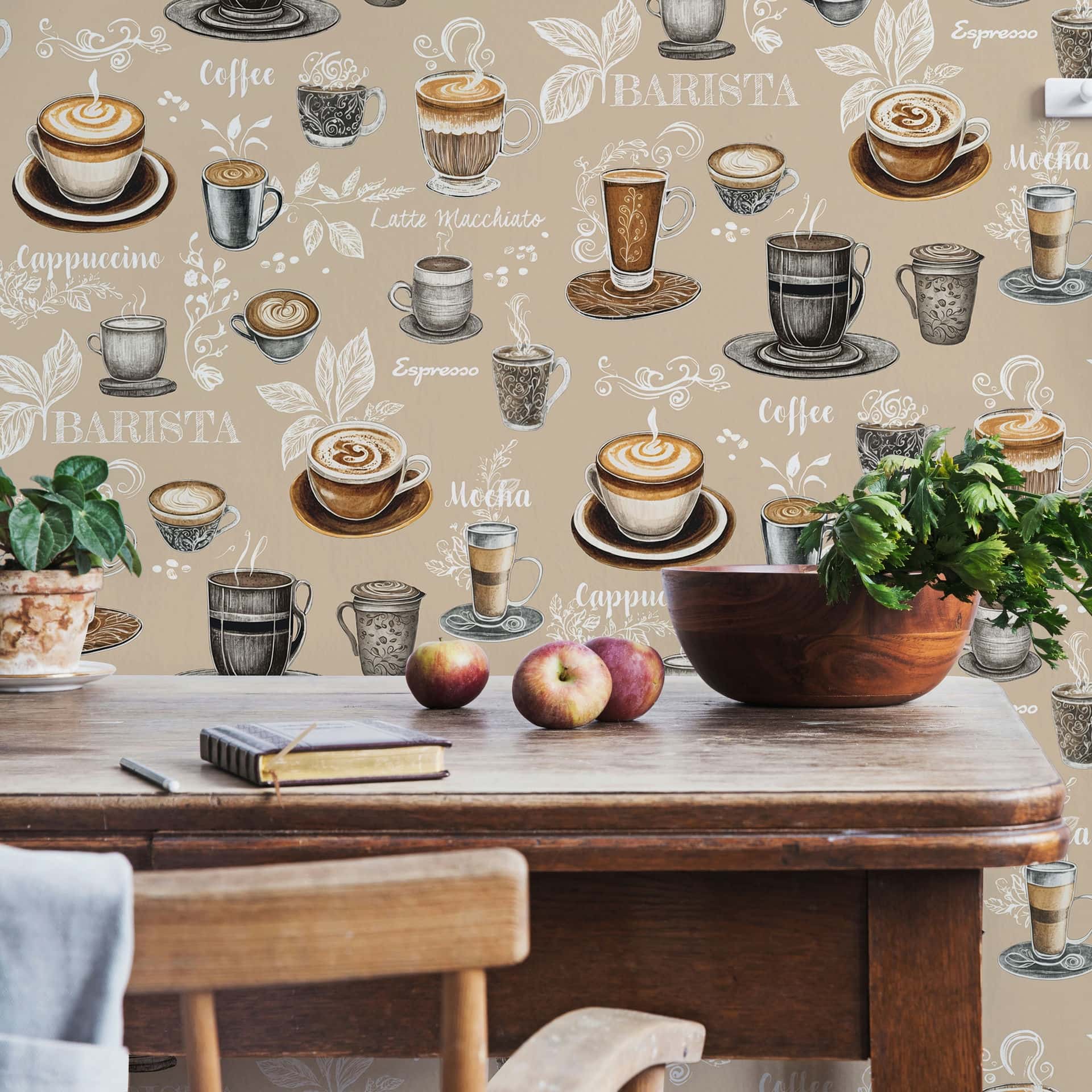Marburg Naomi 89044 Beige wallpaper with illustrated coffee cups, mugs, saucers, and coffee names forms the backdrop to a wooden dining table with a chair, apples, a wooden bowl, a potted plant, and a notebook with a pencil.