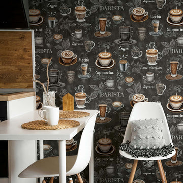 Marburg Naomi 89045 Coffee-themed wallpaper on a dark background with illustrated cups, saucers, leaves, and hand-lettered coffee names behind a small kitchen dining area with a white table, chairs, a mug, and wood-accent cabinets.