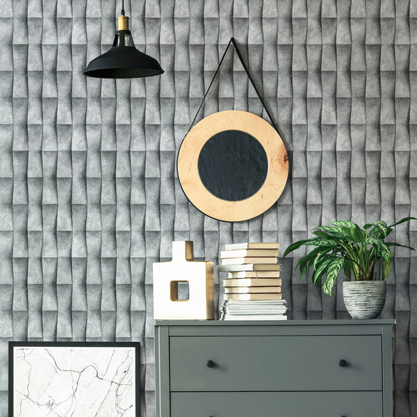 Marburg Novamur Mona 81649 Gray 3D‑effect geometric wallpaper shown behind a gray dresser styled with stacked books, a sculptural object, a potted plant, a round hanging mirror, a framed print, and a black pendant light.