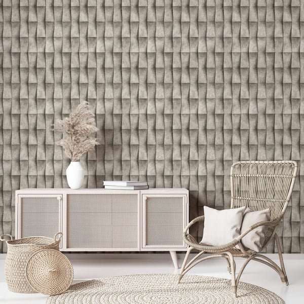 Marburg Novamur Mona 81650 Beige‑grey geometric wallpaper with a 3D folded effect behind a light cane-front sideboard, a white vase with dried grasses and books, a rattan chair with cushions, a woven basket, and a round braided rug.
