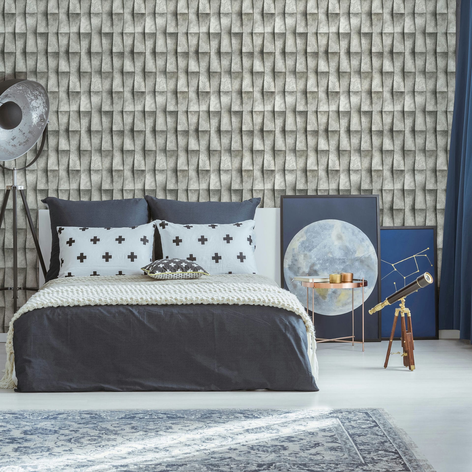 Marburg Novamur Mona 81651 Bedroom scene with a gray 3D geometric patterned wallpaper behind a made bed. A tripod floor lamp stands to the left, framed art leans on the floor beside a round metallic side table, a telescope on a wooden tripod sits to the right, with curtains and a rug in.
