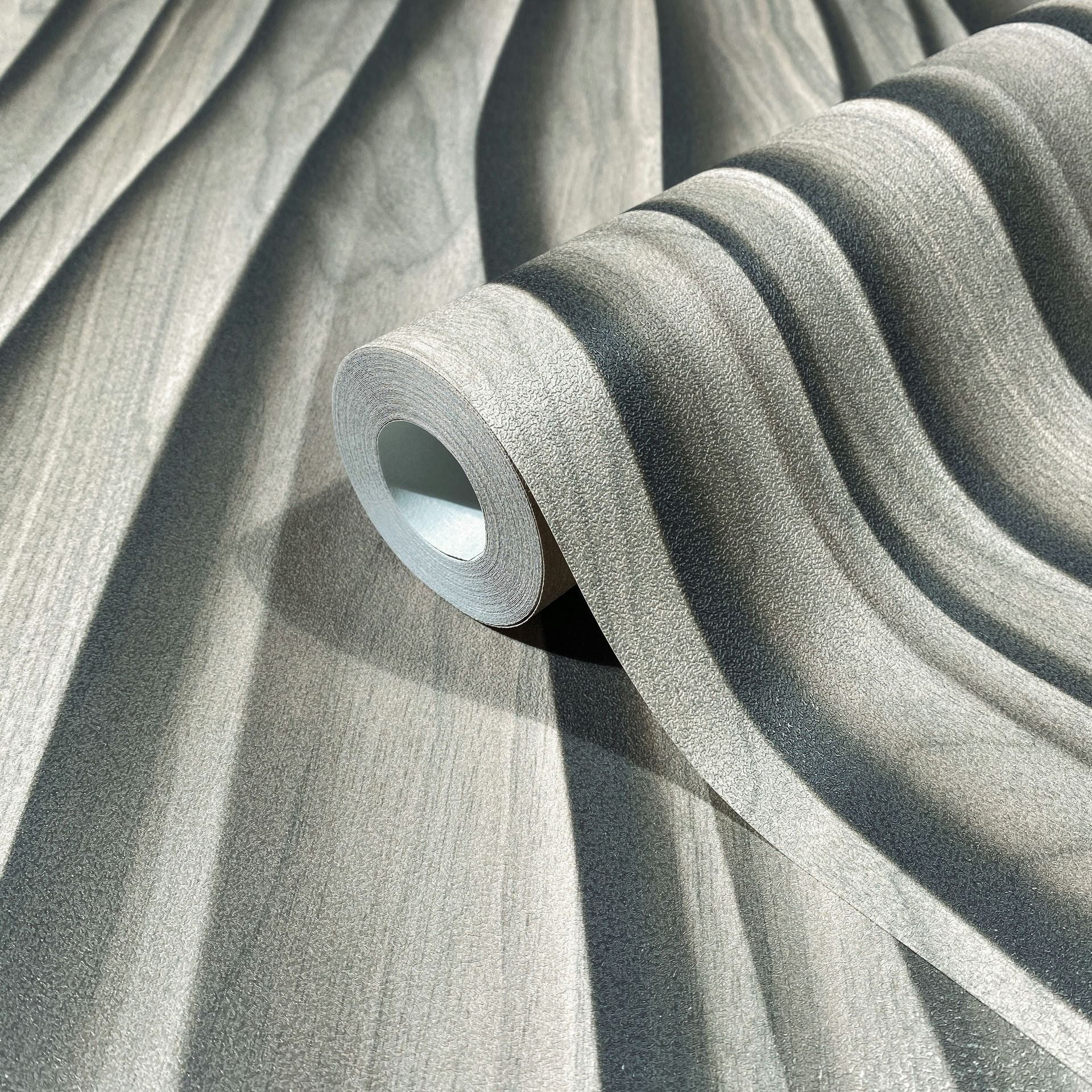 Marburg Novamur Mona 81665 Close-up of a wallpaper roll resting on matching paper, displaying a soft wavy stripe pattern in light beige-gray with a textured surface.