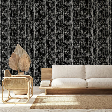 Marburg Novamur Mona 81672 Black wallpaper with a dense bamboo stalk and leaf pattern behind a light wood sofa and a rattan chair with dried leaves, with a natural fiber rug on the floor.