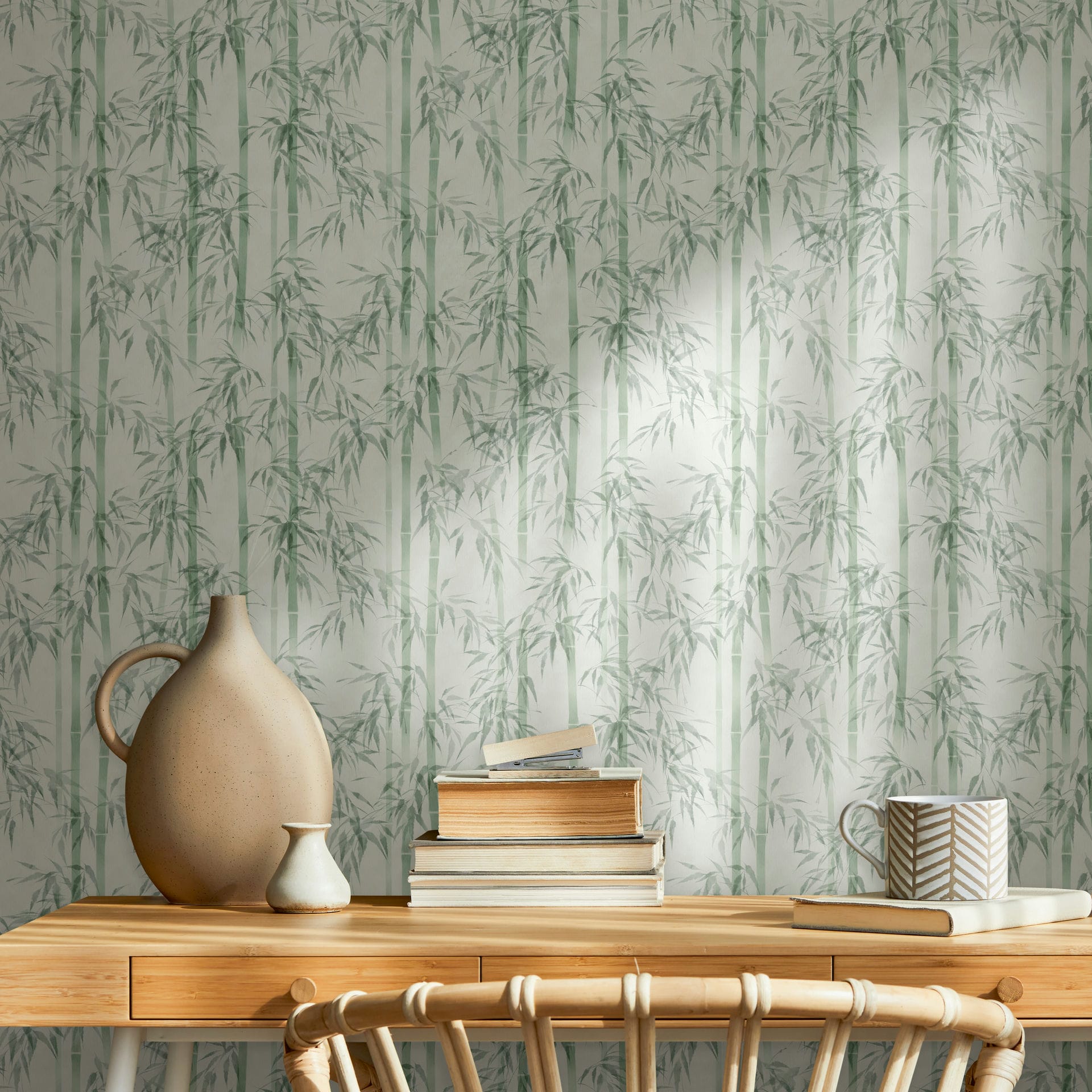 Marburg Novamur Mona 81674 Green bamboo pattern wallpaper behind a wooden desk styled with stacked books, a patterned mug, a large ceramic jug-style vase, a small vase, and a chair.