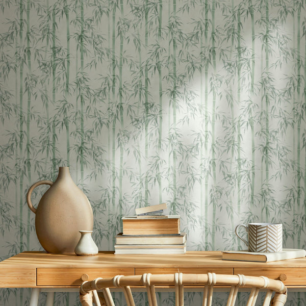 Marburg Novamur Mona 81674 Green bamboo pattern wallpaper behind a wooden desk styled with stacked books, a patterned mug, a large ceramic jug-style vase, a small vase, and a chair.