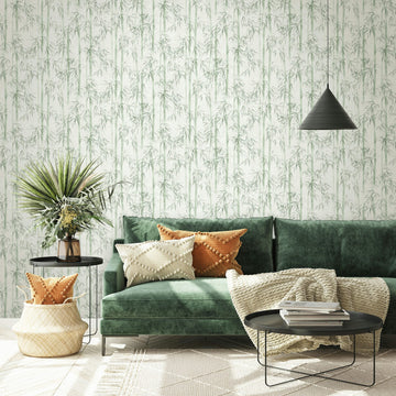 Marburg Novamur Mona 81674 Living room scene featuring green bamboo pattern wallpaper on a light background behind a green sofa, round black tables, a plant, basket with cushions, a knitted throw, stacked books, and a black cone pendant light.