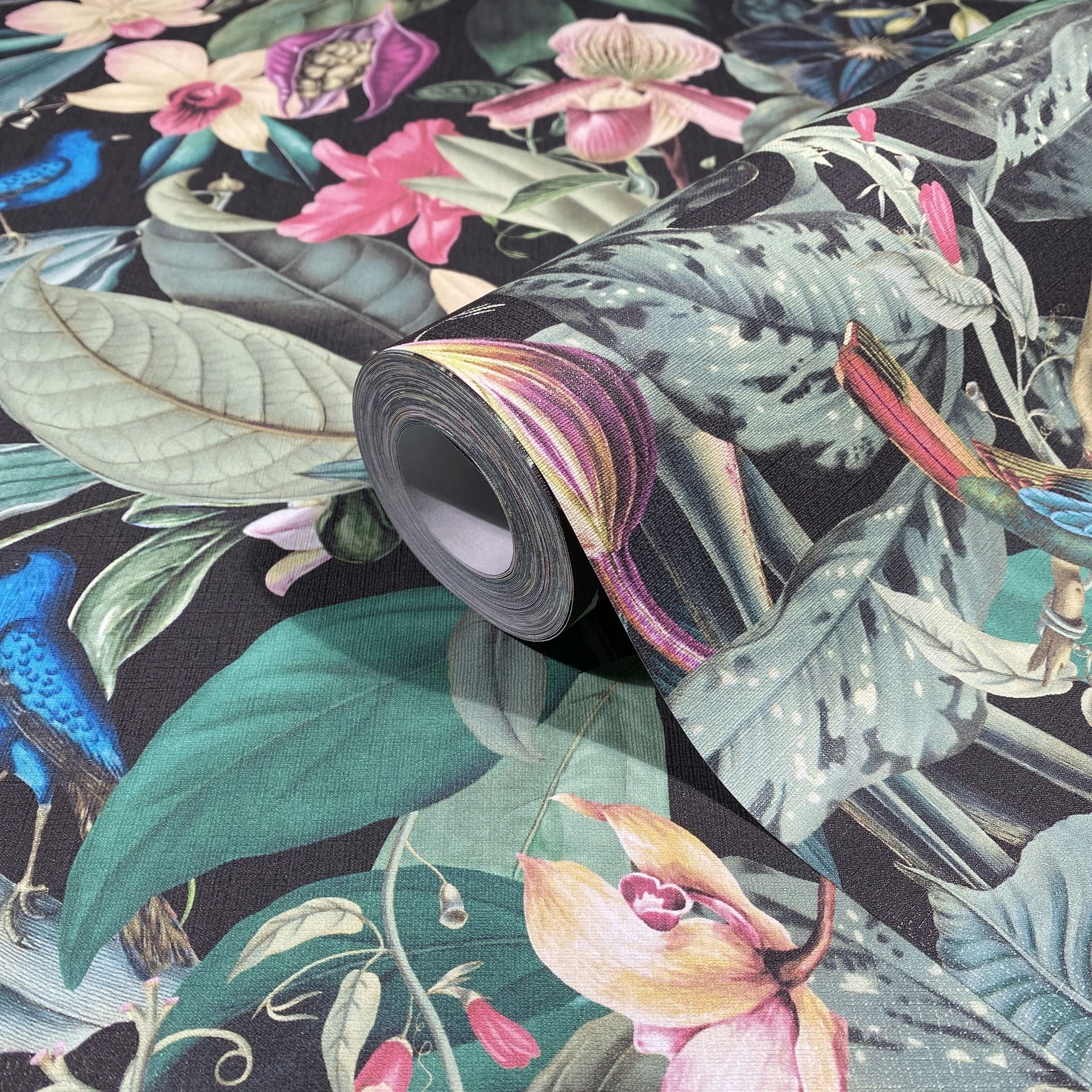 Marburg Novamur Mona 81685 Close-up of a wallpaper roll resting on matching paper, showing a dark-background tropical botanical design with large leaves, colorful flowers, and birds.