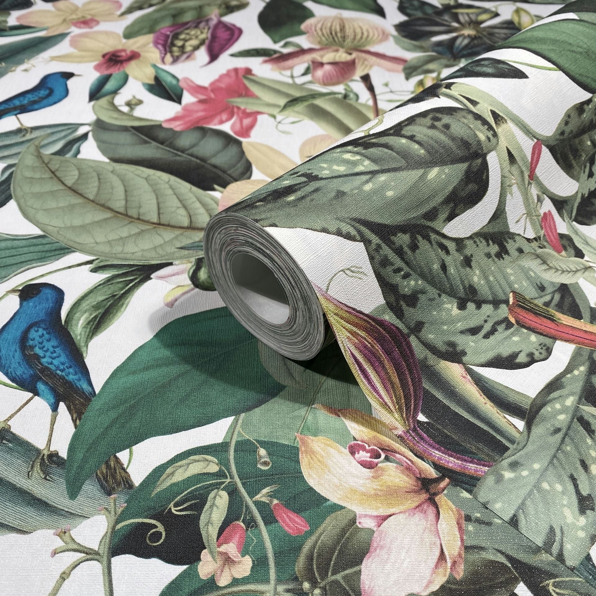 Marburg Novamur Mona 81686 Angled close-up of a wallpaper roll on matching paper, showing a tropical botanical design with large green leaves, pink and cream flowers, and blue birds on a white background.