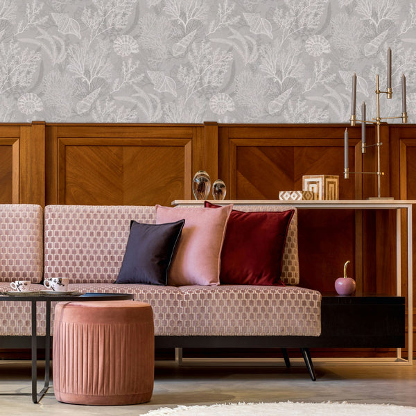 Marburg Novamur Mona 81687 Gray wallpaper with white coral and shell motifs above wood paneling, shown behind a patterned sofa with cushions, a small coffee table with cups, a round ottoman, and a console with candlesticks.