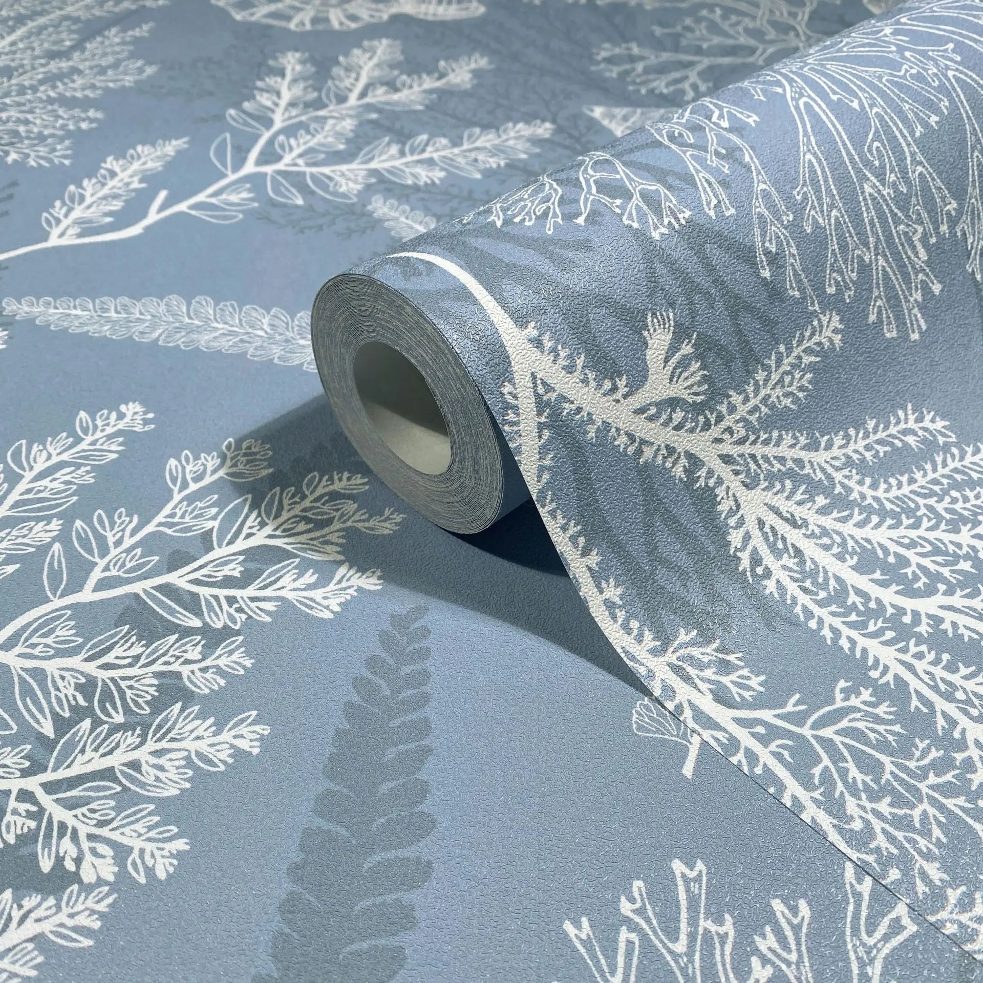 Marburg Novamur Mona 81689 A close-up of a wallpaper roll partly unrolled on matching material, showing white botanical sprigs and fern-like fronds on a muted blue, lightly textured background.