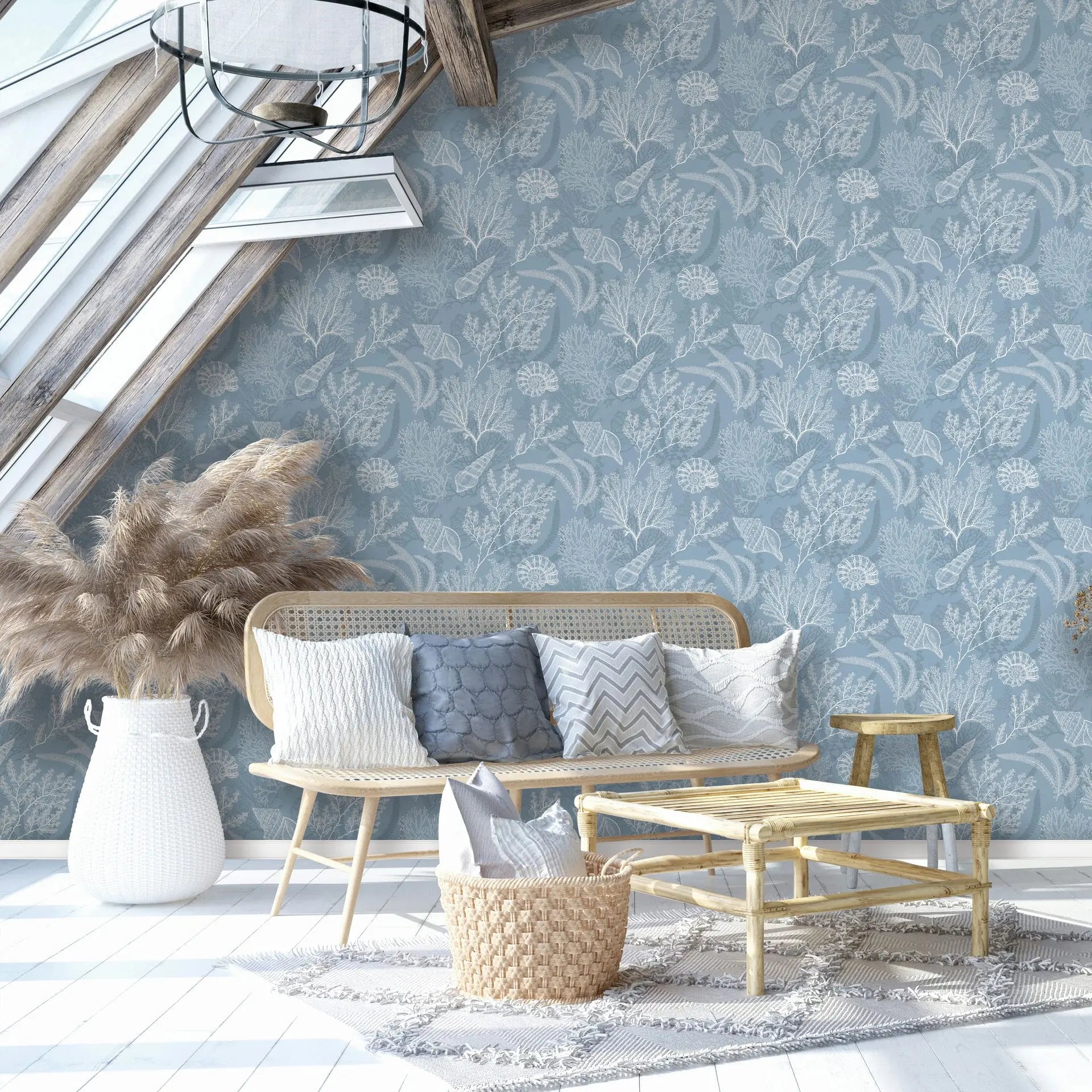 Marburg Novamur Mona 81689 Blue wallpaper with a white coral and seashell pattern is used on a feature wall in a bright attic living room, shown with a rattan sofa, bamboo coffee table, woven basket with pillows, a white woven floor basket holding pampas grass, a wooden stool, rug, skyl.