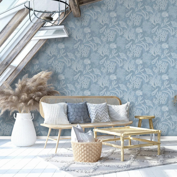 Marburg Novamur Mona 81689 Blue wallpaper with a white coral and seashell pattern is used on a feature wall in a bright attic living room, shown with a rattan sofa, bamboo coffee table, woven basket with pillows, a white woven floor basket holding pampas grass, a wooden stool, rug, skyl.