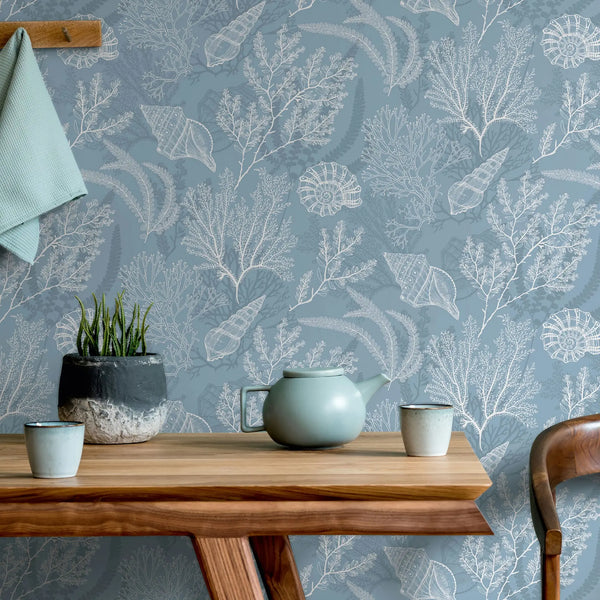 Marburg Novamur Mona 81689 Blue wallpaper with white coral and seashell illustrations is shown behind a wooden table holding a ceramic teapot, two cups, and a small potted plant; a towel hangs on a wooden peg rail and part of a chair is visible.