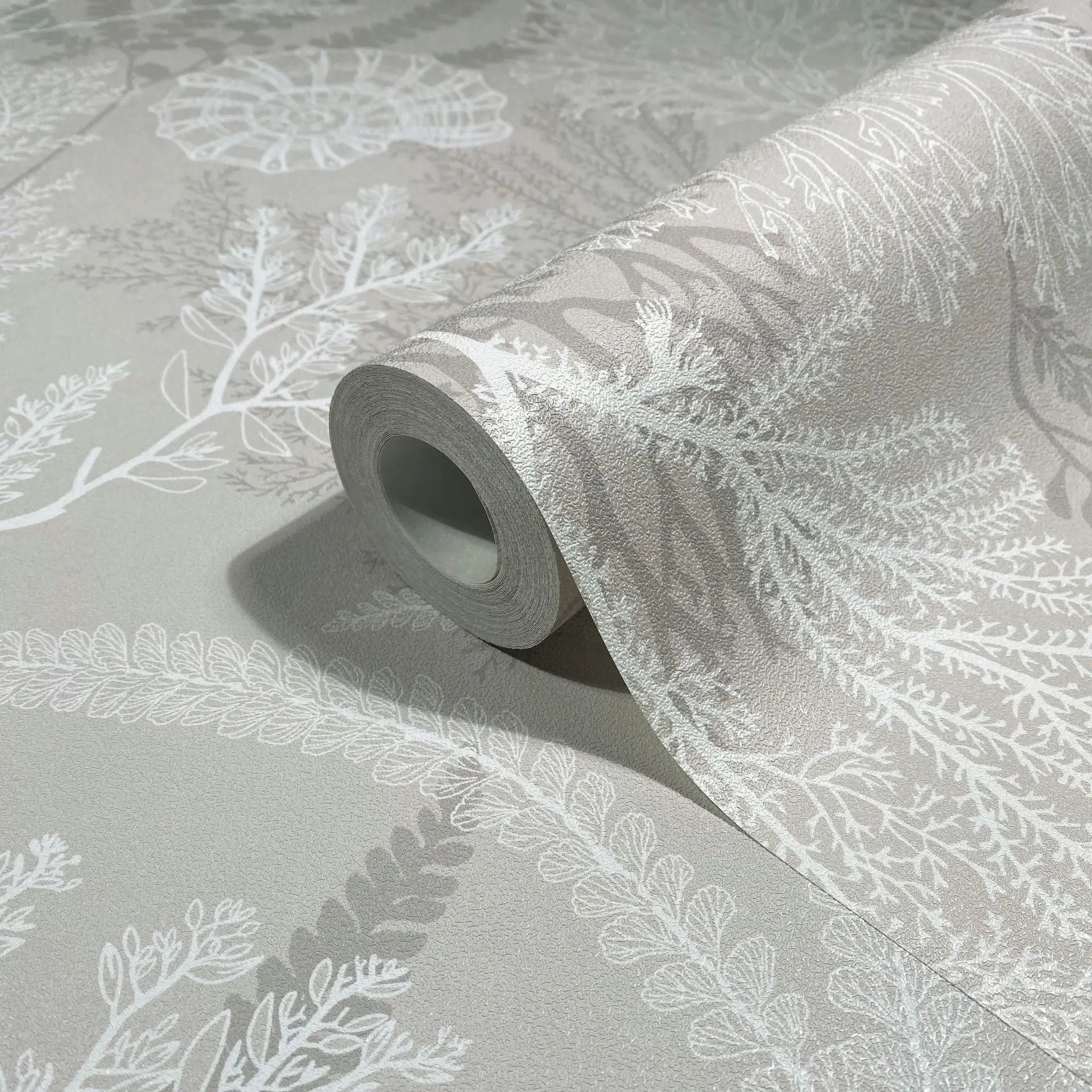 Marburg Novamur Mona 81690 Close-up of a partially unrolled wallpaper roll on a matching surface, showing a beige background with white botanical fronds and leaf motifs.