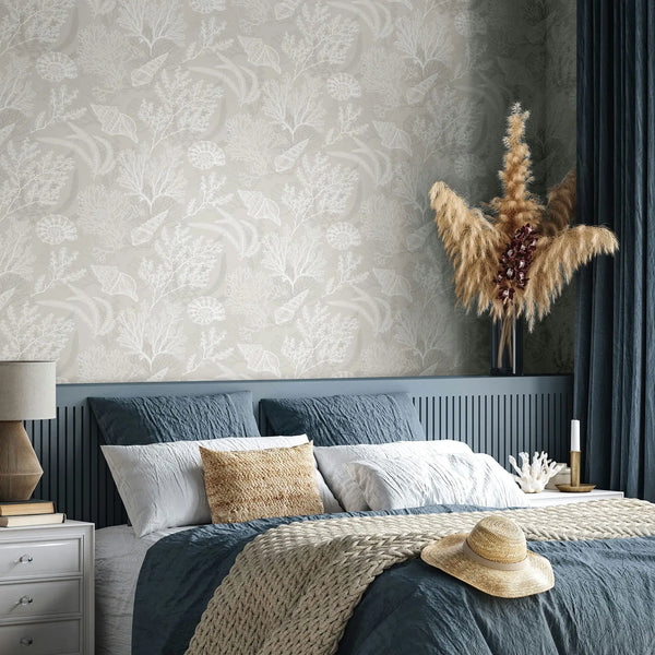 Marburg Novamur Mona 81690 Bedroom scene featuring beige wallpaper patterned with white coral, leaves, and shell motifs behind a bed with blue bedding. A nightstand with a table lamp sits on the left, and on the right are dark curtains, a tall vase of pampas grass, and decor items on a侧.