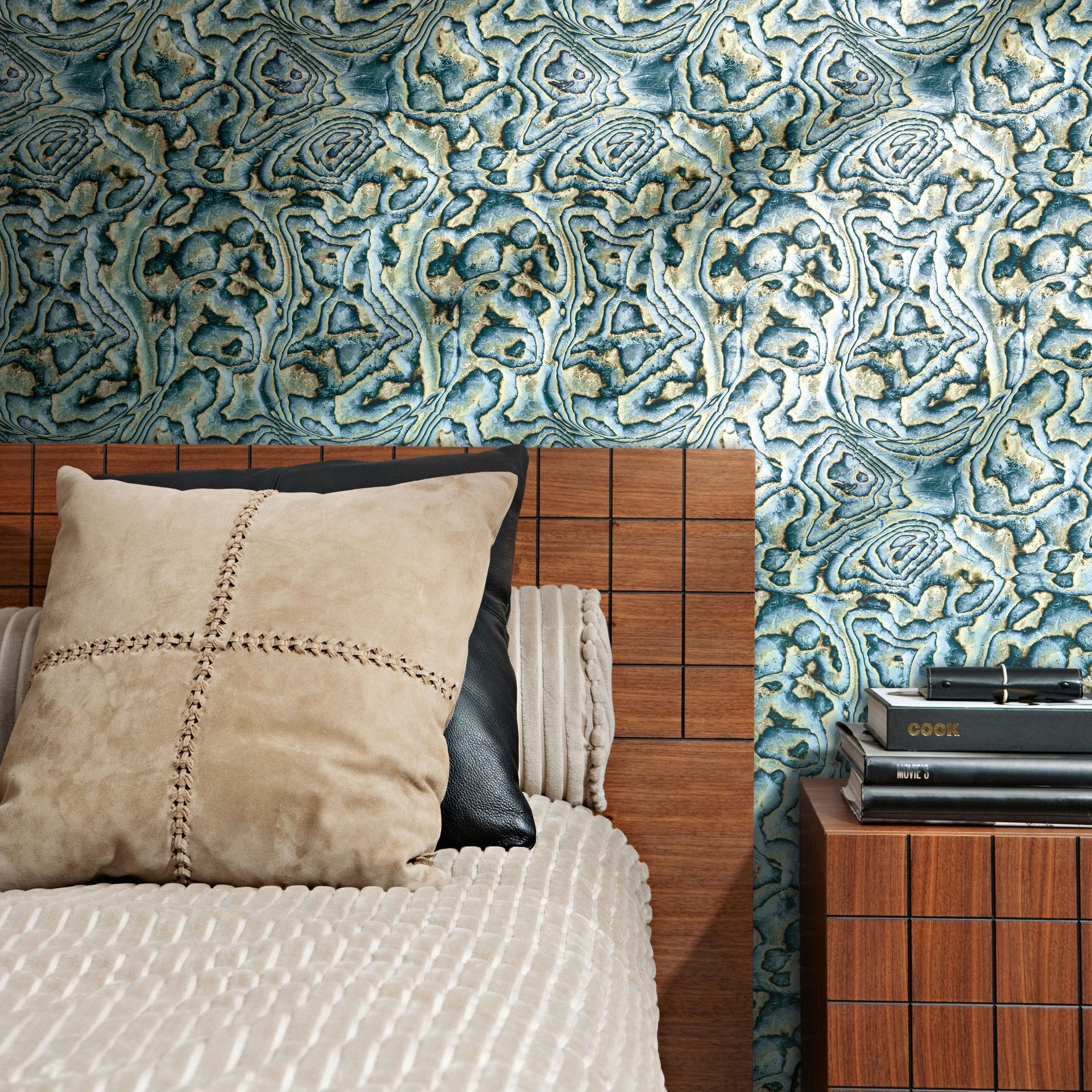 Marburg Novamur Mona 81692 Abstract agate-style wallpaper in blue, green, and beige behind a wood headboard with bedding and a nightstand topped with stacked books.