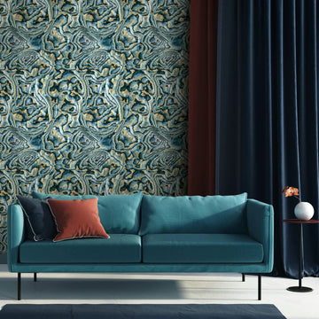 Marburg Novamur Mona 81692 Abstract geode-style wallpaper in blue, green, and beige behind a teal sofa with cushions, dark curtains, and a small side table holding a round vase with a single flower.