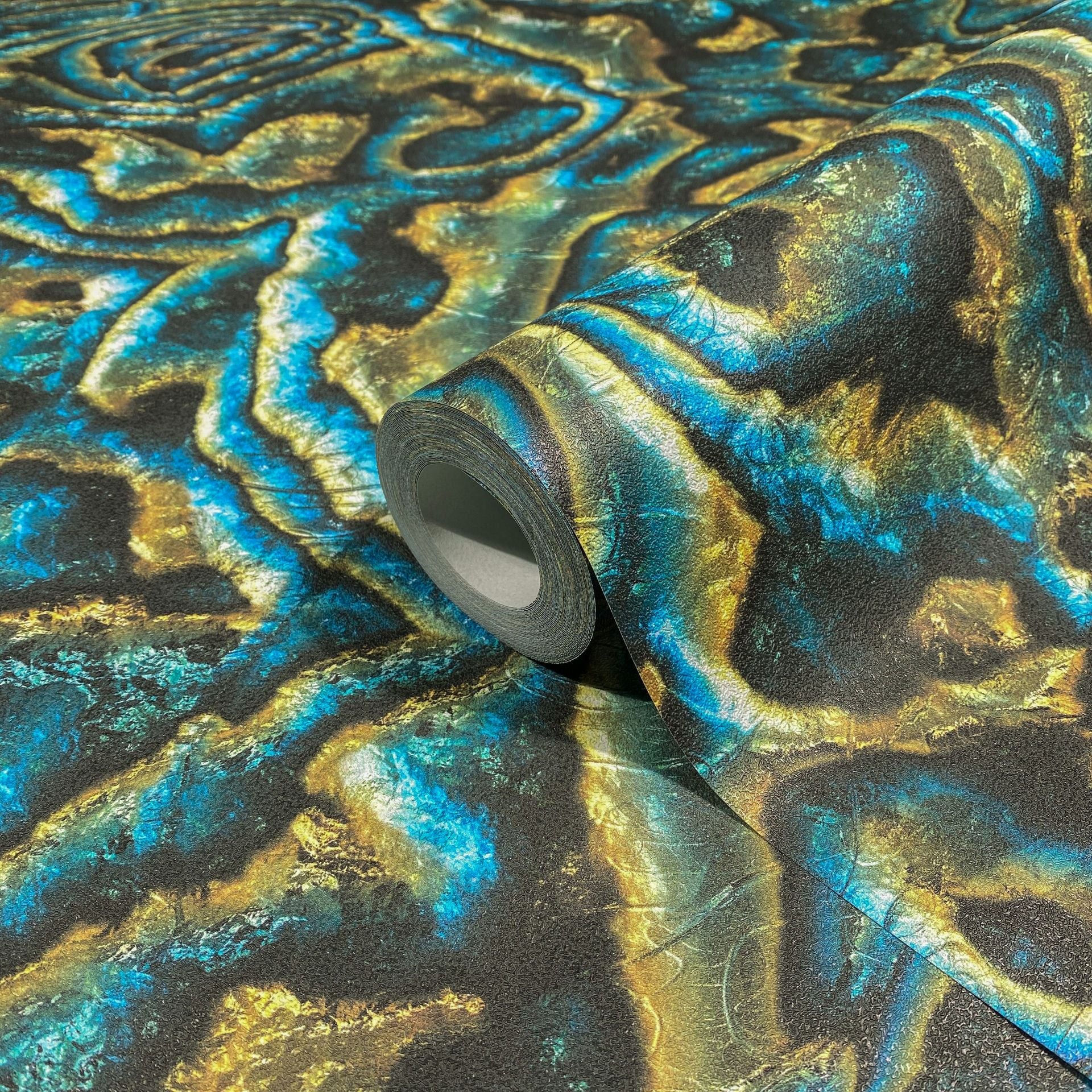 Marburg Novamur Mona 81693 Wallpaper roll resting on matching paper with an abstract blue, teal, and gold marbled pattern; close-up product view showing textured surface.