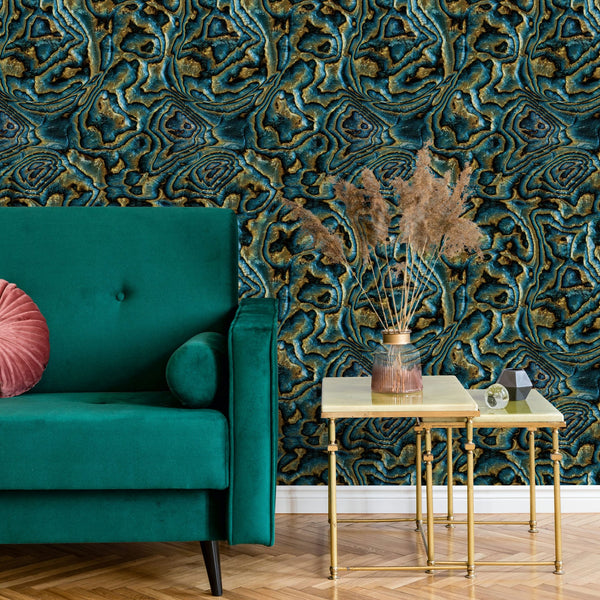 Marburg Novamur Mona 81693 Abstract geode-style wallpaper in blue, teal, and brown behind a teal sofa with a round pink pillow and metal nesting side tables holding a glass vase with dried stems.