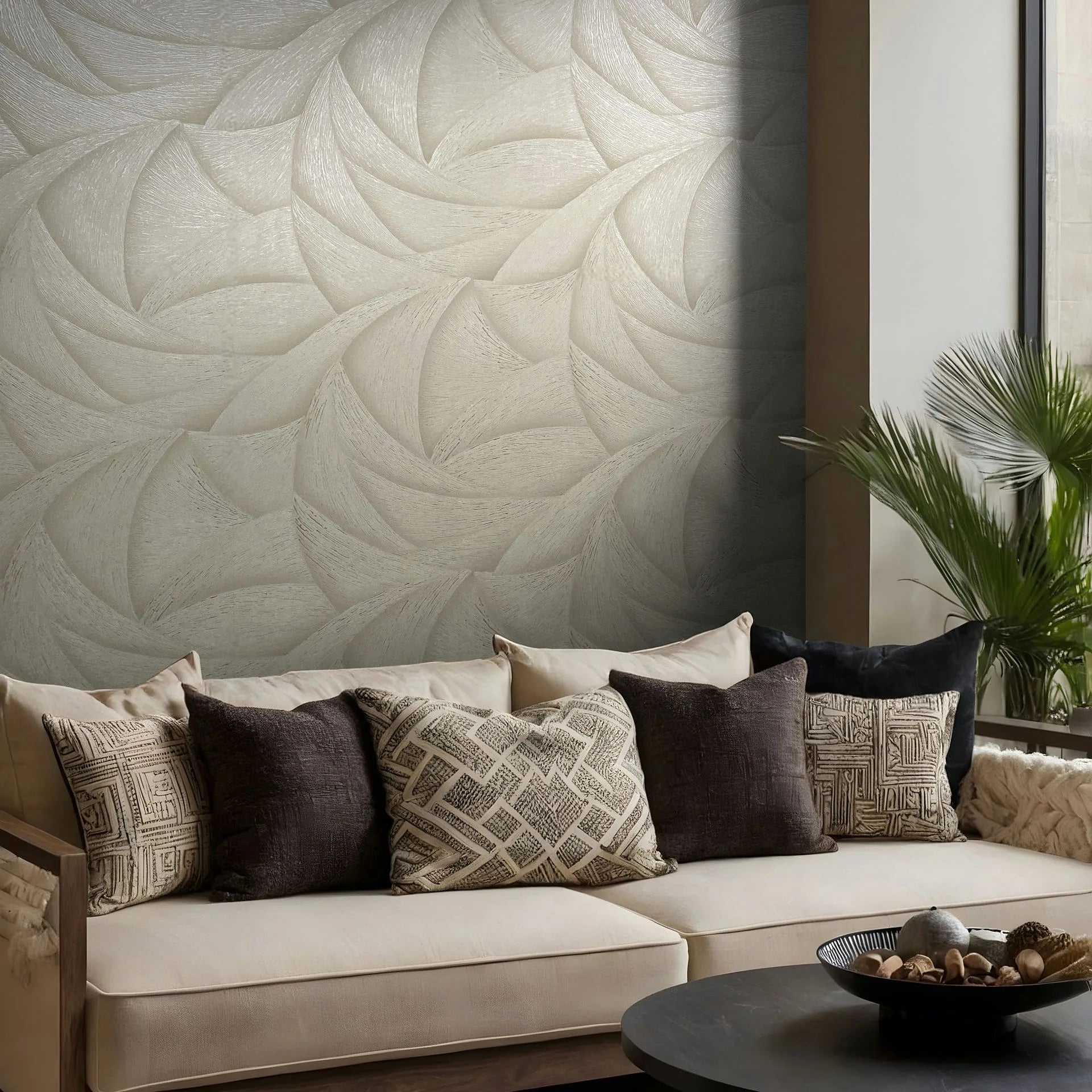 Marburg Ritus 35502 Living room scene with beige wallpaper of overlapping curved leaf-like swirls behind a neutral sofa, with patterned pillows, a round coffee table holding a decorative bowl, and a potted palm.