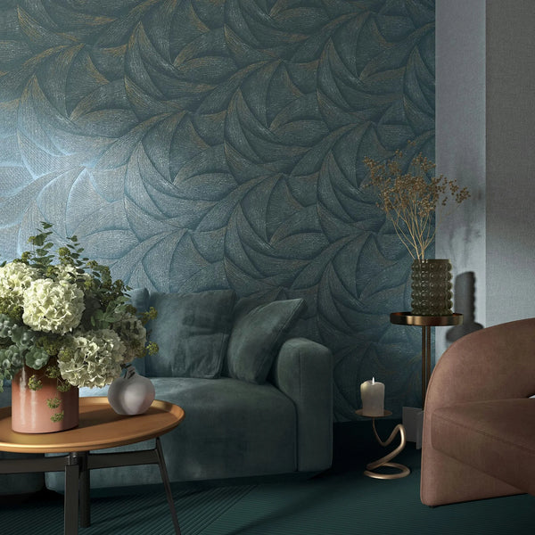 Marburg Ritus 35507 Blue wallpaper with a large, overlapping leaf-like motif and a subtle metallic sheen, shown in a living room with a sofa, a round coffee table holding flowers, a side table with a textured glass vase and dried stems, and a candle on a sculptural holder.