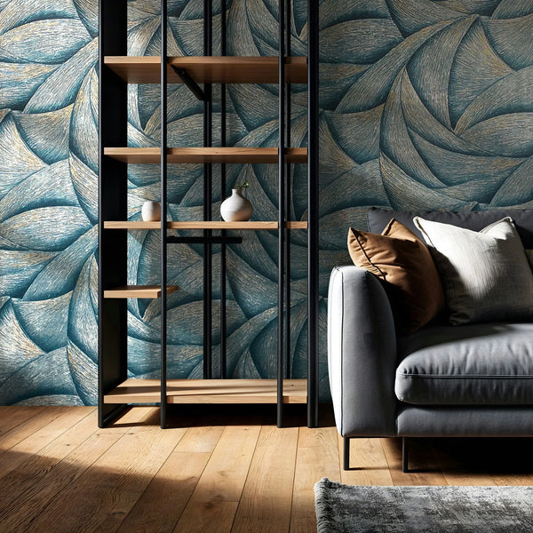 Marburg Ritus 35507 Blue wallpaper with large overlapping leaf-like curves and fine striations, highlighted with light gold tones. A black metal and wood shelving unit and a gray sofa with cushions sit on a wooden floor with a small rug edge visible.