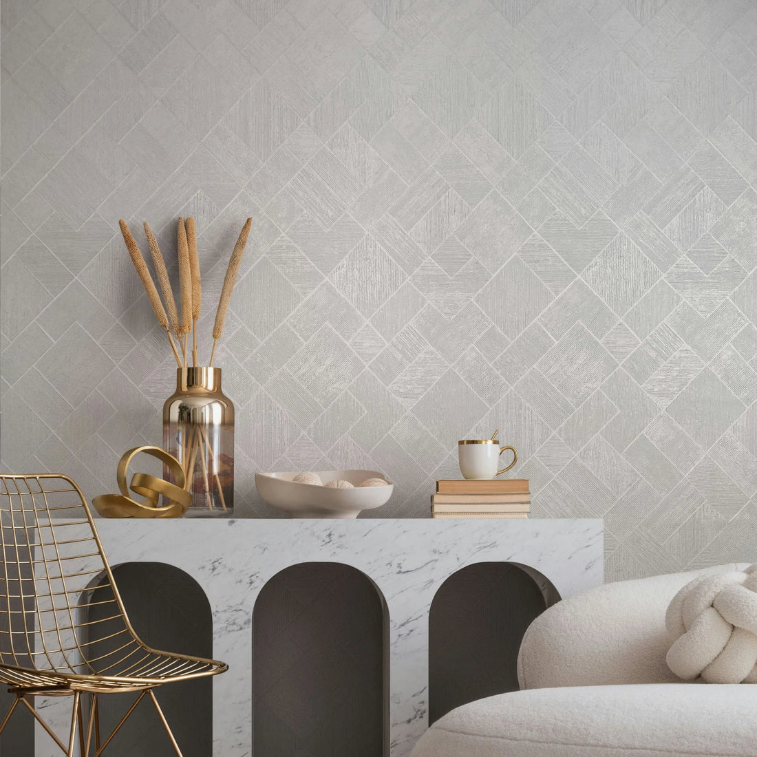 Marburg Ritus 35508 Light neutral geometric wallpaper of small diagonal squares shown behind a marble console styled with a metallic vase holding dried stems, a decorative ring sculpture, a bowl, books with a cup, and a gold wire chair beside a cream sofa.