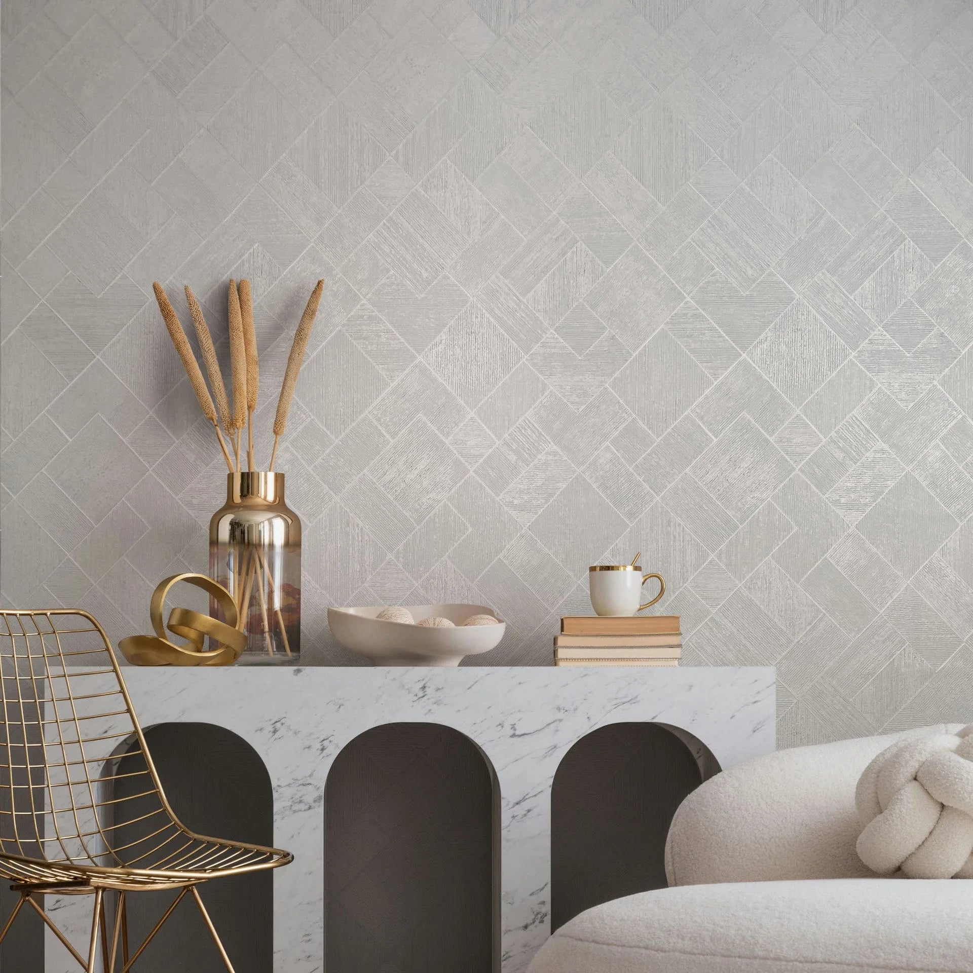 Marburg Ritus 35508 Light neutral geometric wallpaper of small diagonal squares shown behind a marble console styled with a metallic vase holding dried stems, a decorative ring sculpture, a bowl, books with a cup, and a gold wire chair beside a cream sofa.
