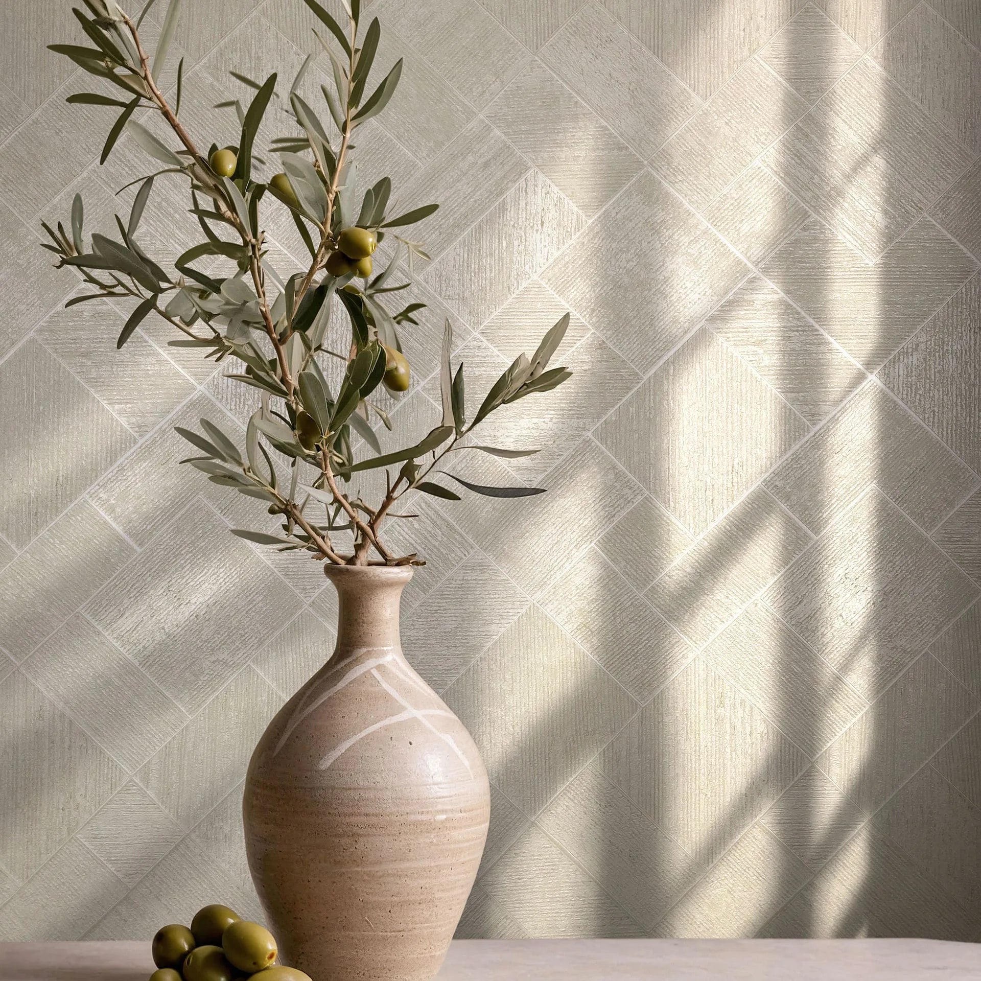 Marburg Ritus 35509 Beige geometric wallpaper with a diagonal parquet of textured squares and diamonds, shown behind a ceramic vase holding an olive branch on a tabletop with soft sunlight casting shadows.