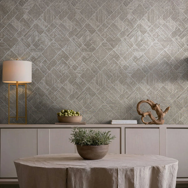 Marburg Ritus 35510 Beige and gray geometric diamond-pattern wallpaper behind a styled dining vignette with a round draped table holding a potted plant, a sideboard with a lamp, books, a wooden sculpture, and a bowl of green fruit.