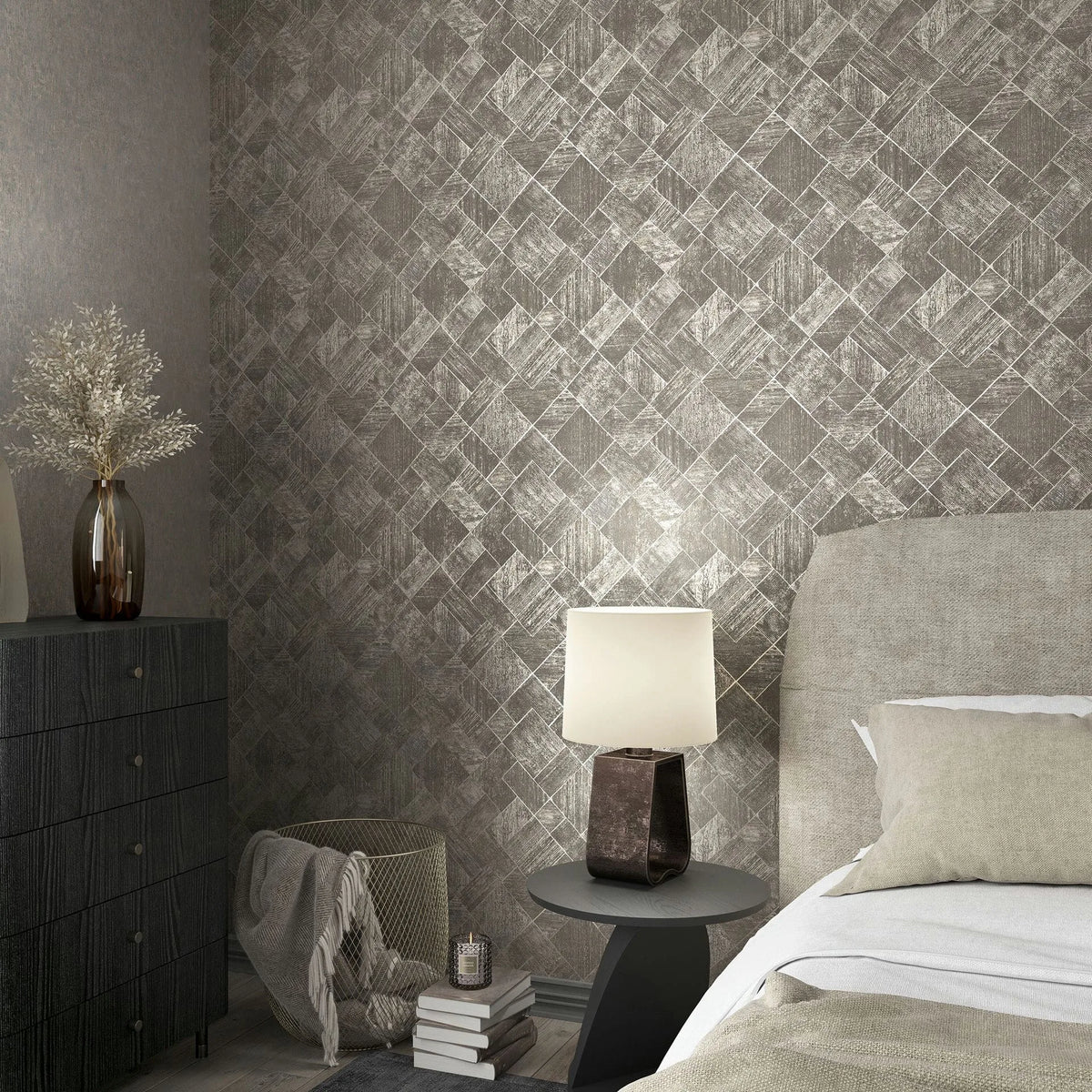 Marburg Ritus 35511 Bedroom scene featuring gray and silver geometric diamond wallpaper. An upholstered bed with neutral bedding sits beside a round side table with a lamp, next to a dark dresser holding a glass vase of dried stems; a basket with a blanket and stacked books is in.