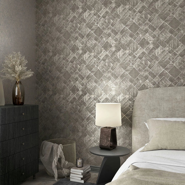 Marburg Ritus 35511 Bedroom scene featuring gray and silver geometric diamond wallpaper. An upholstered bed with neutral bedding sits beside a round side table with a lamp, next to a dark dresser holding a glass vase of dried stems; a basket with a blanket and stacked books is in.