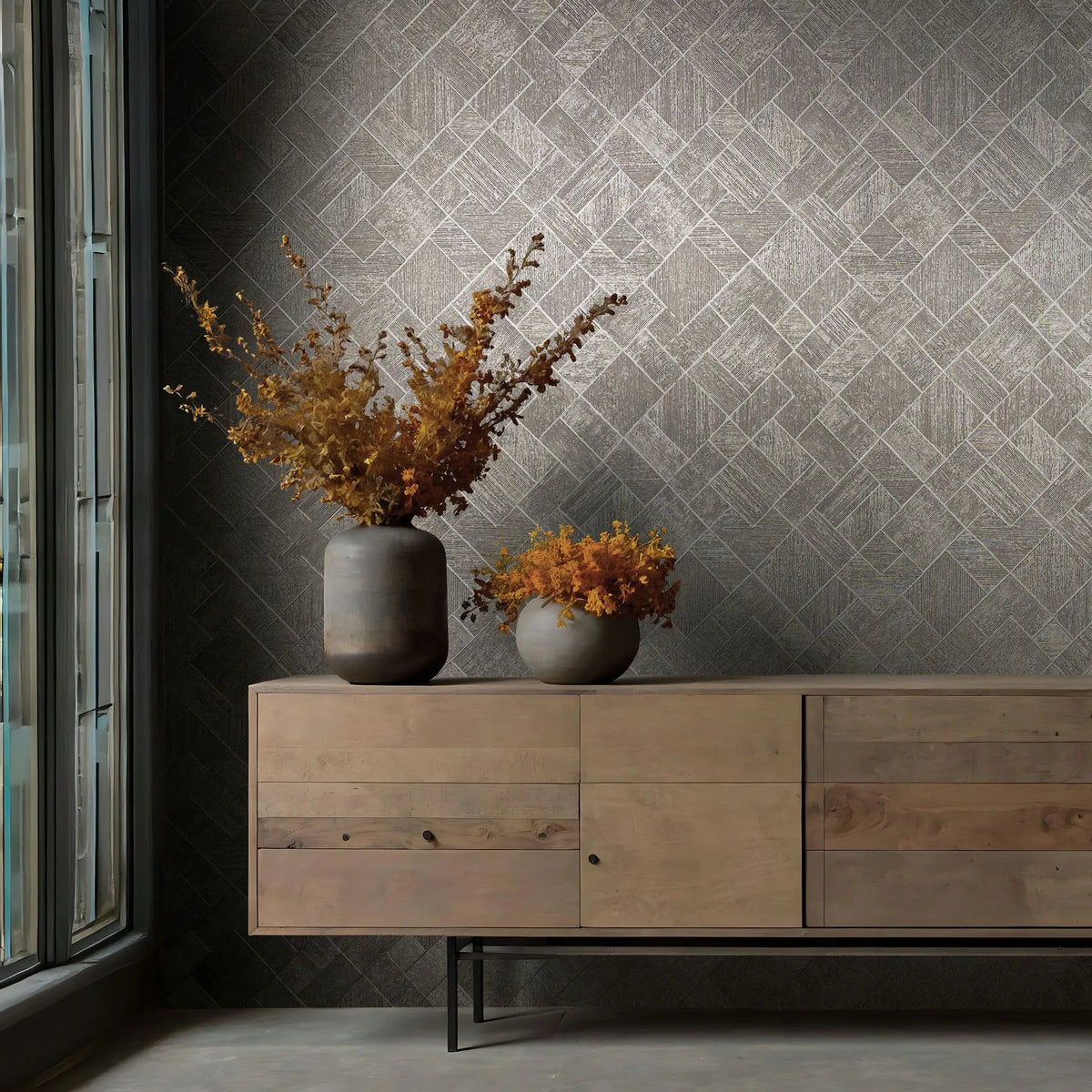 Marburg Ritus 35511 Gray geometric wallpaper with a diamond grid of small rectangular blocks, shown with a wooden sideboard, two vases holding dried foliage, and a window on the left.