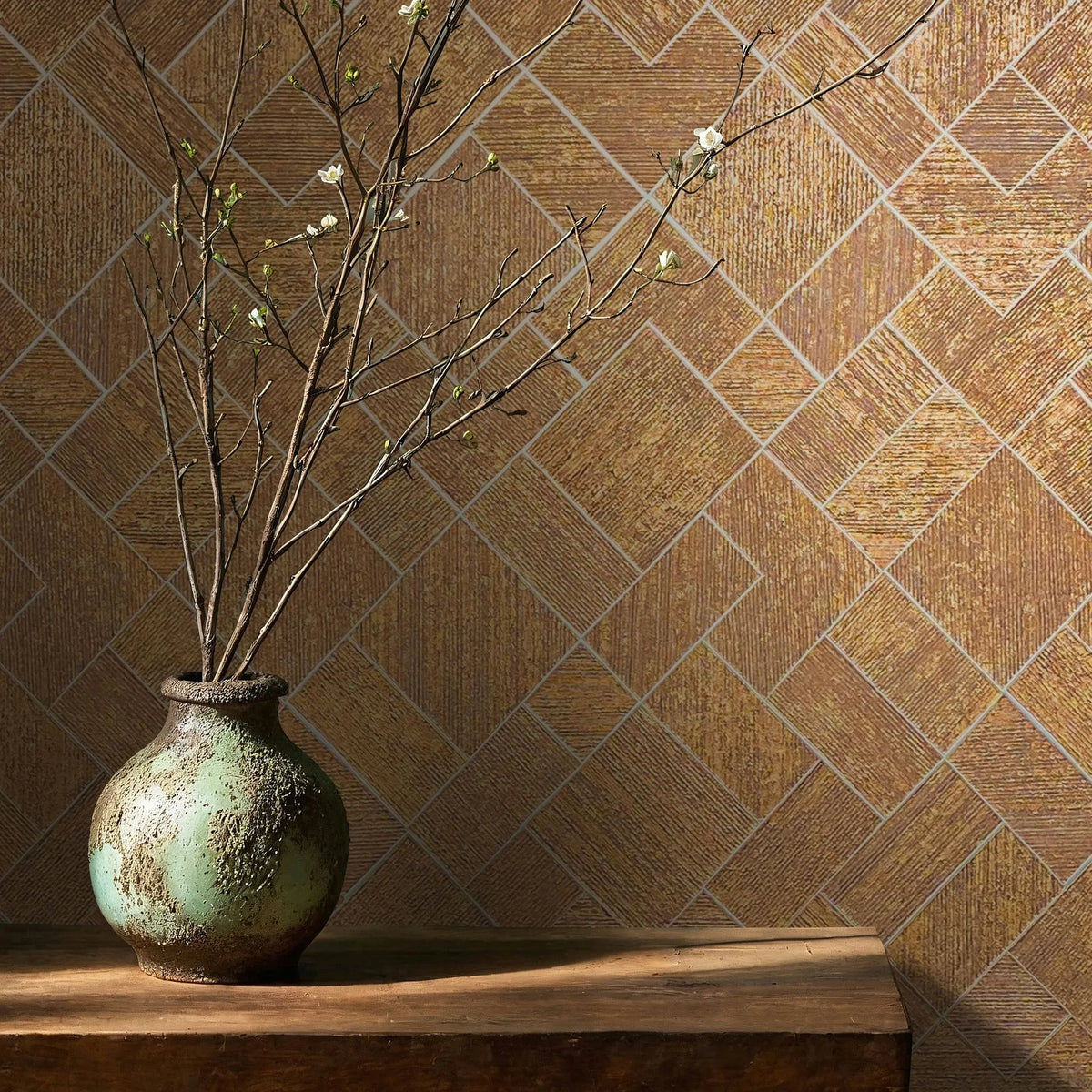 Marburg Ritus 35513 Geometric wallpaper of diagonal rectangular blocks in warm gold tones, shown with a rustic ceramic vase of branches on a wooden table.