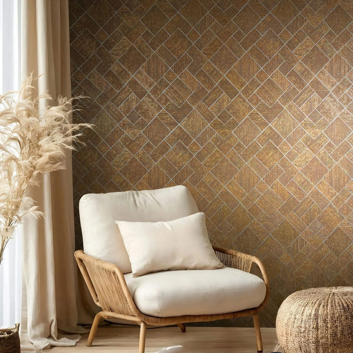 Marburg Ritus 35513 Gold-toned geometric herringbone-style wallpaper behind a rattan armchair with a cream cushion and pillow, a woven pouf, beige curtains, and pampas grass.