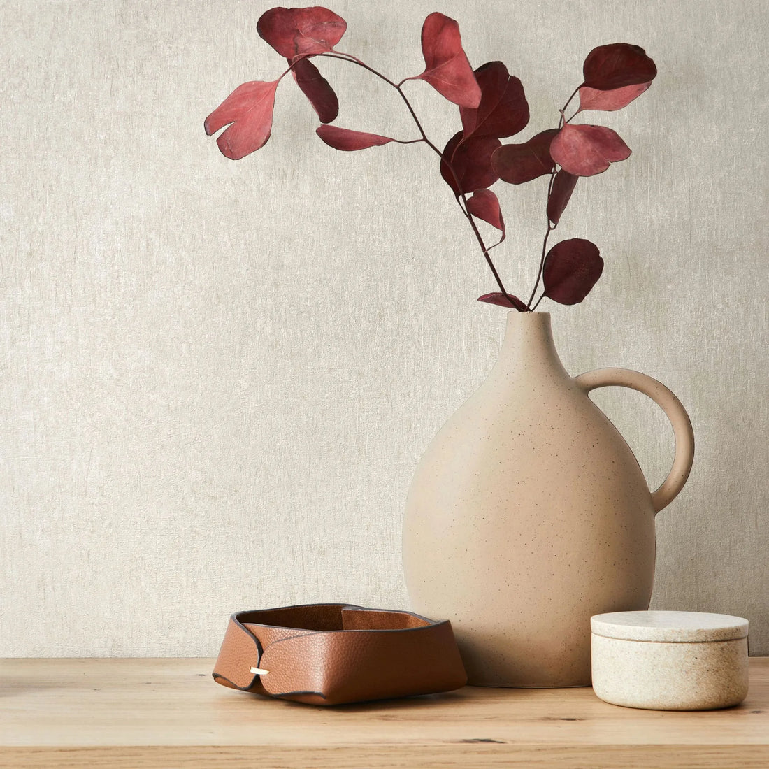 Marburg Ritus 35522 Light beige textured wallpaper behind a matte ceramic jug holding red leaves, with a small leather tray and a round lidded container on a wooden surface.