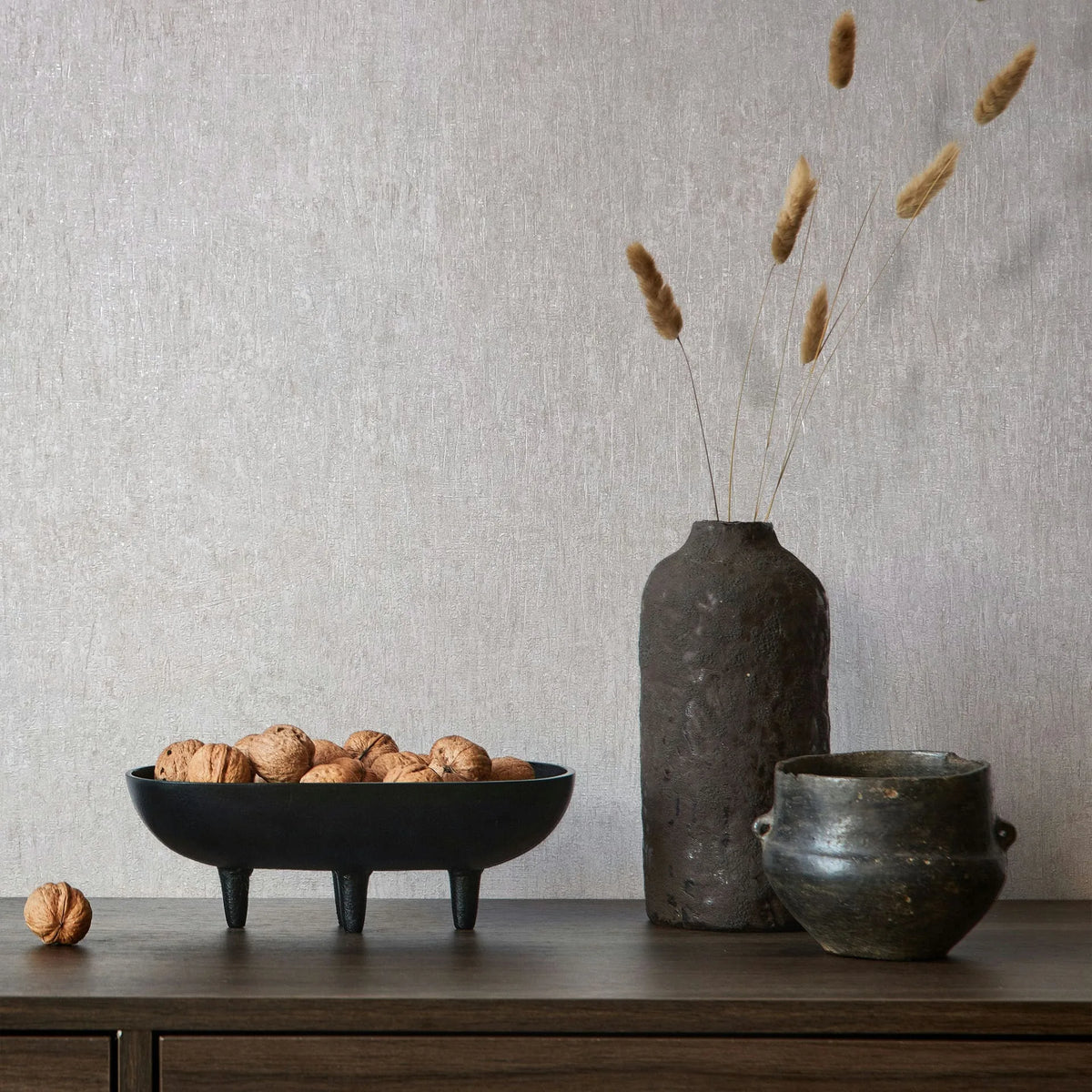 Marburg Ritus 35523 Light gray textured wallpaper behind a wood sideboard with a black three-legged bowl filled with walnuts, a tall dark vase holding dried grasses, and a small pot.