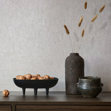 Marburg Ritus 35523 Light gray textured wallpaper behind a wood sideboard with a black three-legged bowl filled with walnuts, a tall dark vase holding dried grasses, and a small pot.