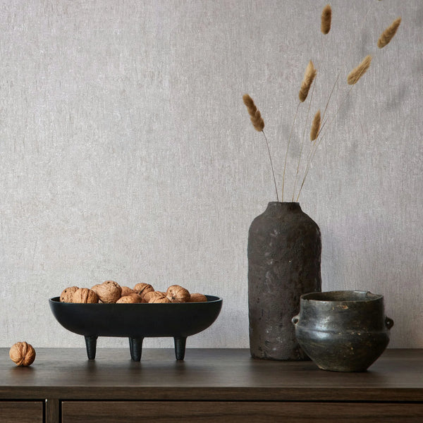 Marburg Ritus 35523 Light gray textured wallpaper behind a wood sideboard with a black three-legged bowl filled with walnuts, a tall dark vase holding dried grasses, and a small pot.