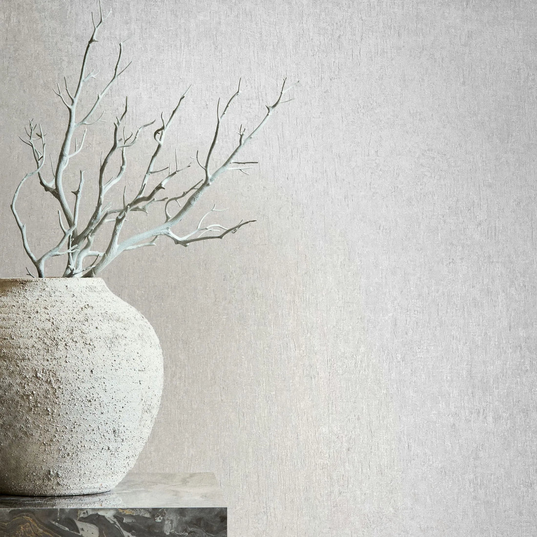 Marburg Ritus 35524 Light neutral textured wallpaper shown behind a rough ceramic pot holding bare branches on a stone pedestal.