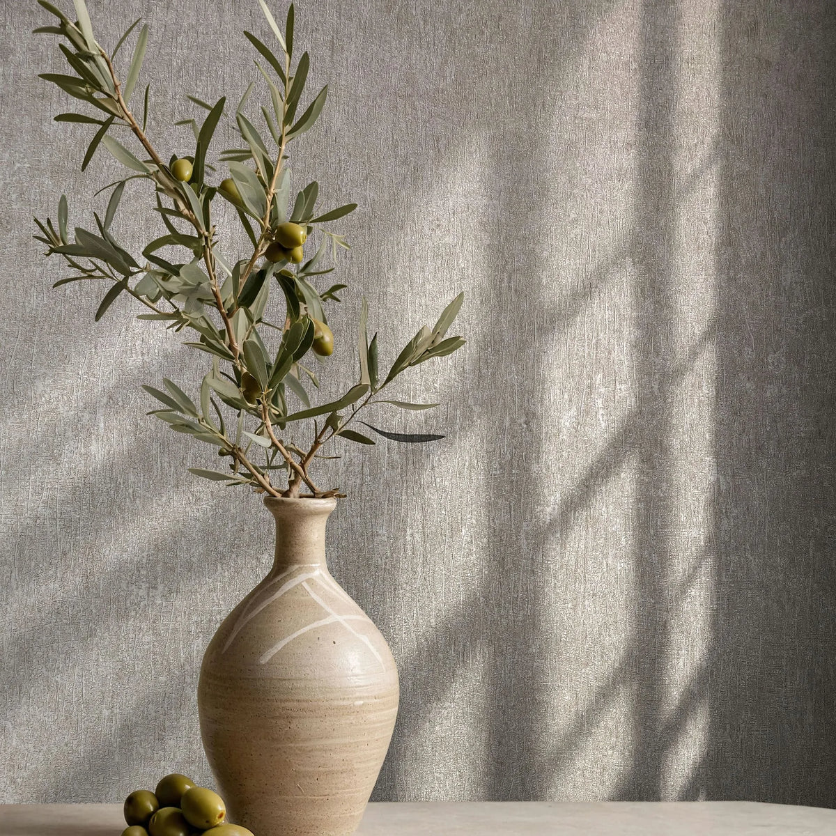 Marburg Ritus 35525 Gray textured wallpaper with a subtle crosshatch pattern, shown behind a ceramic vase holding olive branches with a few loose olives on a tabletop and window-light shadows on the wall.
